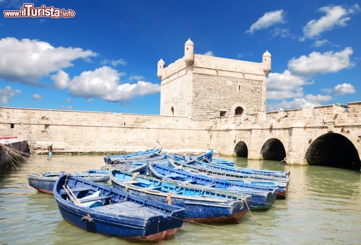 Immagine Forte e porto di Essaouira, Marocco - Secondo la tradizione questa città che si affaccia sull'Oceano Atlantico fu fondata da mercanti cartaginesi in un luogo abitato da popolazioni berbere. Diventata presto un importante scalo commerciale sulla rotta verso il golfo di Guinea, attorno al III° secolo a.C. venne riconquistata dai berberi che vi instaurarono una monarchia. In questa immagine il porto peschereccio di Essaouira con le sue barche dalle mille tonalità azzurre ritratto con il forte sullo sfondo © Waj / Shutterstock.com