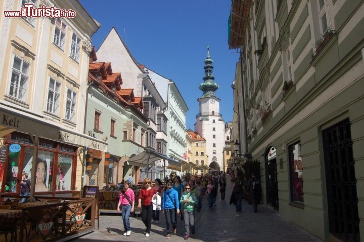 Immagine Via Michalska a Bratislava, Slovacchia - Cittadini e turisti passeggiano lungo la centrale via Michalska, nel cuore della capitale slovacca, su cui si affacciano negozi e ristoranti. Sullo sfondo si intravede la Torre di San Michele che oggi ospita al suo interno un museo di armi antiche: dalla sua sommità, a 51 metri di altezza, si gode di un panorama eccezionale sul cuore della città.