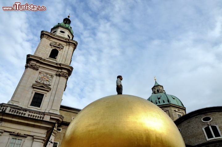 Immagine La Sfera di Salisburgo, in Kapitelplatz, ritratta con il Duomo della città sullo sfondo. Alta circa 9 metri compreso il piedistallo, questa creazione artistica rappresenta una figura maschile in piedi in posa rilassata posizionata su una sfera d'oro.