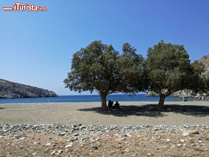 Immagine Spiaggia di Vatses, isola si Astypalaia (Grecia), con persone all'ombra di un albero. E' un ampia baia di ciottoli con scogliere ai lati e una distesa di mare azzurro in mezzo.