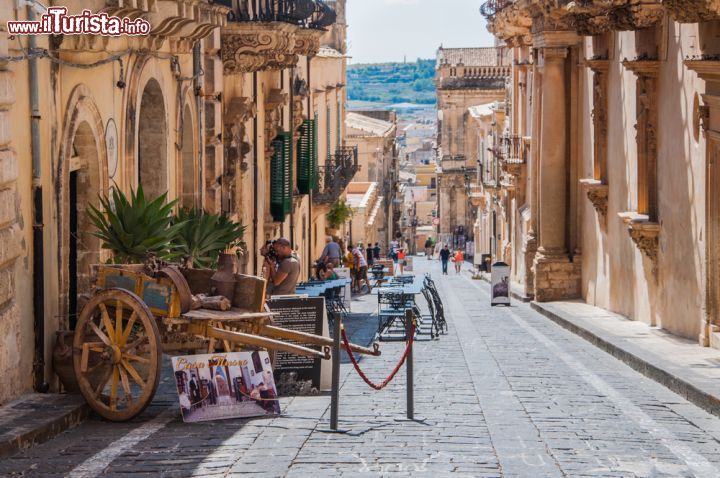 Immagine Cartoline di Sicilia: Noto - il carretto siciliano, simbolo del folclore contadino di questa bella regione, apre a una delle vie principali del centro storico di Noto, denominato "patrimonio dell'umanità" dall'UNESCO, per il suo essere un vero tripudio del barocco siciliano. - © Frog Dares / Shutterstock.com
