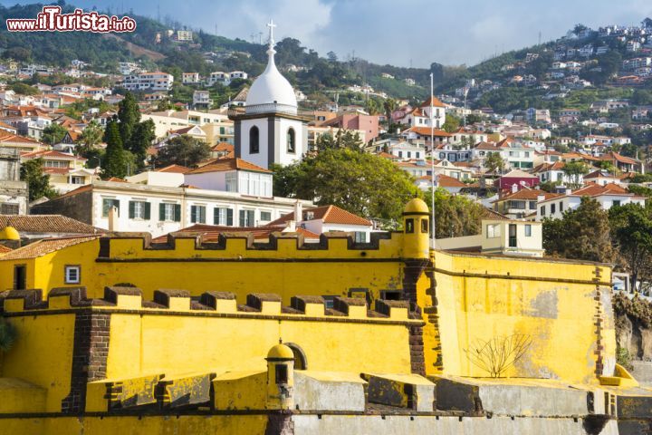 Immagine Il castello Fortaleza de Sao Tiago a Funchal, Madeira (Portogallo) - Nell'immaginario collettivo un castello è imponente, fiabesco e per alcuni versi anche potenzialmente terrificante, ha colori scuri e le porte di legno scricchiolano, nascondendo al loro interno chissà quale mistero o leggenda. A Madera no e a Funchal ancora meno. La Forteza de Sao Tiago ha una tinta piuttosto insolita, un bel giallo limone, mentre le sue torri/decorazioni merlettate (e da questo si capisce che il complesso aveva funzioni militari) come si può vedere dall'immagine, sono marrone scuro. Un colpo d'occhio particolare guardato insieme ai tetti di tutta la città - © Alberto Loyo / Shutterstock.com