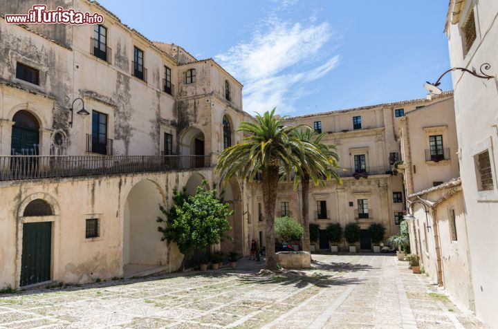 Immagine Cortile del Palazzo Nicolaci di Villadorata: il palazzo dei principi - è una cartolina tipica siciliana quella che possiamo ammirare in questa foto; dove le palme donano colore alla pietra bianca e rosata degli edifici, e dove il barocco regna sovrano. Il Palazzo, che ora ospita in una parte la biblioteca comunale di Noto, fu fatto costruire verso la metà del XVIII secolo come dimora nobiliare urbana della prestigiosa famiglia Nicolaci, i principi di Villadorata.- © LYSVIK PHOTOS / Shutterstock.com