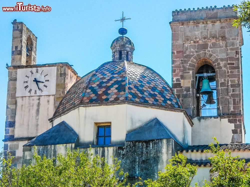 Immagine Dettagli della chiesa dell'Immacolata Concezione nel centro di Barumini, Sardegna: una torre campanaria con l'orologio, la cupola colorata e il campanile. Le forme tardogotiche sono riconoscibili soprattutto nel campanile a canna quadrata.