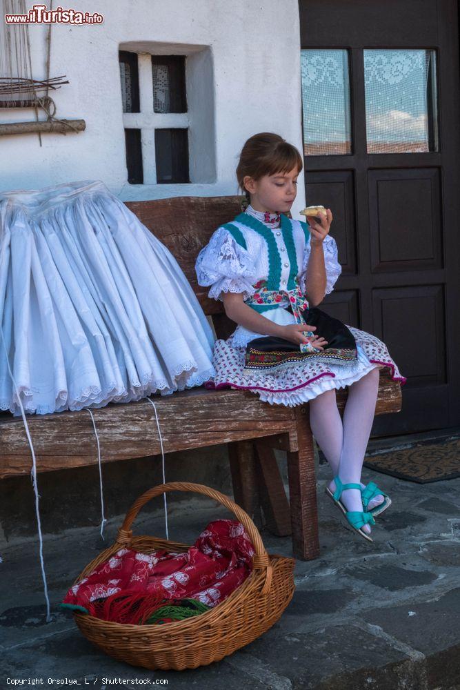 Immagine Holloko, Ungheria: una bambina con abiti tradizionali seduta su una panca davanti a una casa del villaggio - © Orsolya_ L / Shutterstock.com