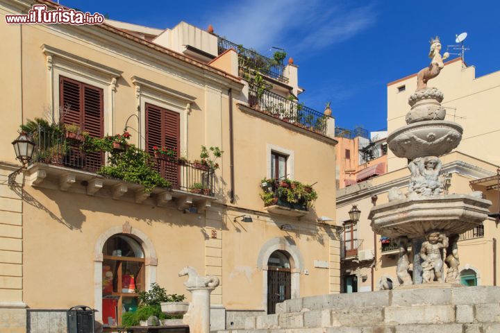 Immagine La fontana di Piazza Duomo con edifici e balconi fioriti sullo sfondo, Taormina. Un caratteristico scorcio fotografico nel cuore della città siciliana.