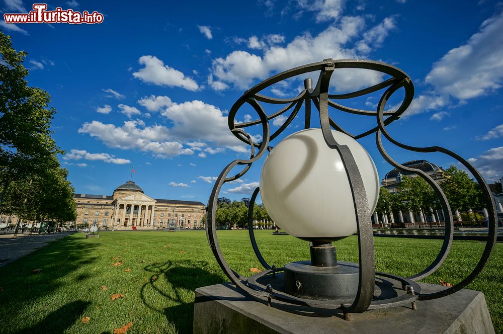 Immagine Una lampada in un giardino di Wiesbaden, Germania, con sullo sfondo l'edificio del Casinò. Una tappa obbligata della città è il Kurhaus, il centro congressi che ospita anche il famoso Casinò Wiesbaden, circondato dal verde e splendidi edifici storici. Il Kurhaus è stato costruito in stile neoclassico ed è sormontato da un’enorme cupola in vetro.