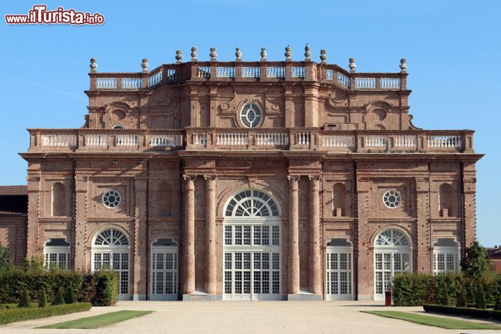 Immagine Vista frontale del Palazzo Venaria Reale, Torino (Piemonte) - Se si guarda in alto sembra la cattedrale di Notre Dame a Parigi, se si guarda il colore sembra il Palazzo Rosso di Genova e se si guarda in basso il Duomo di Bologna. Evidenti le colonne che abbracciano i finestroni centrali, miti i rosoni, pacate le ringhiere che contornano il grandissimo terrazzo, elegantissimo l'ellisse in vetro posto al centro. L'impatto visivo, merito anche di un color terra bruciata di Siena, nell'insieme trasmette fascino e mistero, lasciando lo spettatore ammaliato - © Emituu / Shutterstock.com