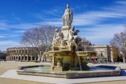 Nimes (Francia) in una giornata di sole con il cielo blu. Al centro dell'immagine, una antica fontana nella piazza antistante l'arena romana.



