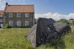 Una tipica barca in legno rovesciata su un prato a Lindisfarne, Inghilterra. Sullo sfondo, eleganti edifici in pietra. 

