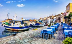 Uno scorcio del porto sull'isola di Chalki, Grecia, con barche ormeggiate e taverne con tavoli e sedie.

