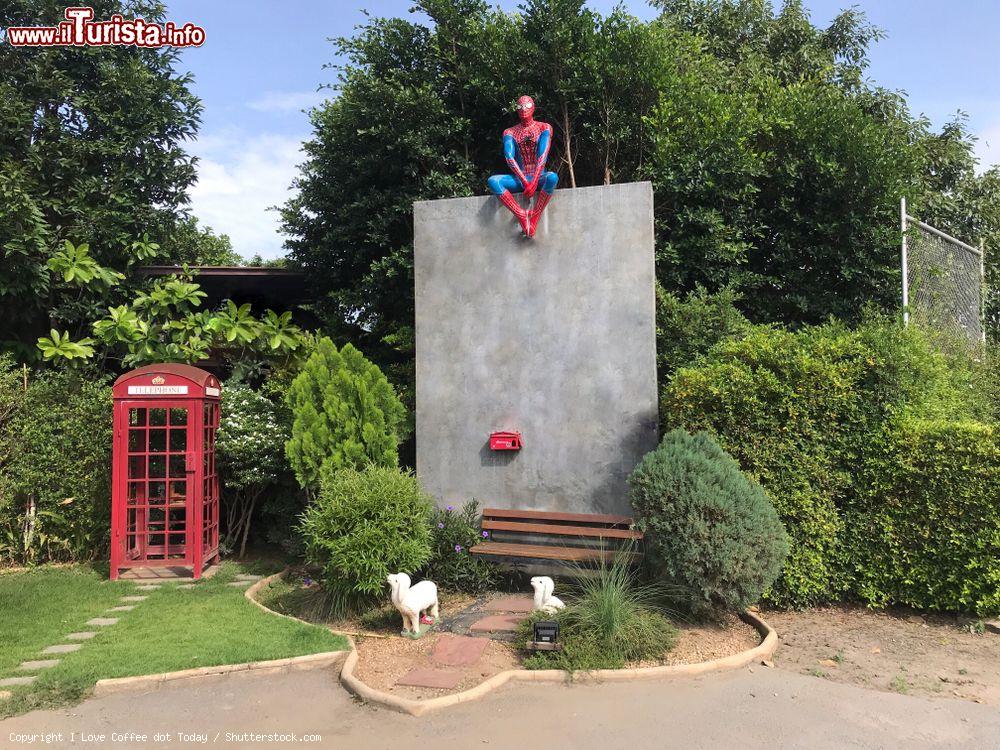 Immagine Statua di Spiderman seduto su un muro nella città di Suphan Buri, Thailandia. Sotto, una panchina con due agnellini e una cabina telefonica rossa - © I Love Coffee dot Today / Shutterstock.com