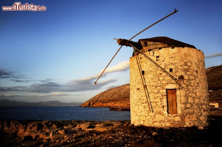 Immagine Il litorale di Kassos (Dodecaneso) al tramonto con il mulino a vento - Romantico, antico e "con quella faccia un po' così" come cantava Paolo Conte riferendosi a Genova, quando un tramonto incontra la spiaggia si vede il paradiso ma, quando nelle luci del crepuscolo si può avere la compagnia di un mulino a vento, allora si comincia a desiderare di non essere morti ma al contrario più vivi che mai. Le sfumature che l'azzurro del cielo riesce ad assumere sono così tante che riflesse non solo sul mare limpido, ma anche sulle pietre granitiche di quello che da lontano sembra essere un granaio in disuso, riescono ad abbracciare e coinvolgere ogni senso - © Antlio / Shutterstock.com