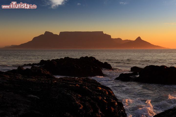 Immagine Tramonto di Table Mountain, Città del Capo (Sudafrica) - Di solito le montagne si trovano a margine di una città, ma non è il caso di Table Mountain. Inglobata nel cuore di Città del Capo, si estende per ben tre chilometri e il suo perimetro dritto, fa apparire il tramonto come una bellissima evocazione quasi mistica, come si può vedere dall'immagine. L'atmosfera soave che si percepisce regala inoltre una scenografia decisamente romantica - © Dietmar Temps / Shutterstock.com