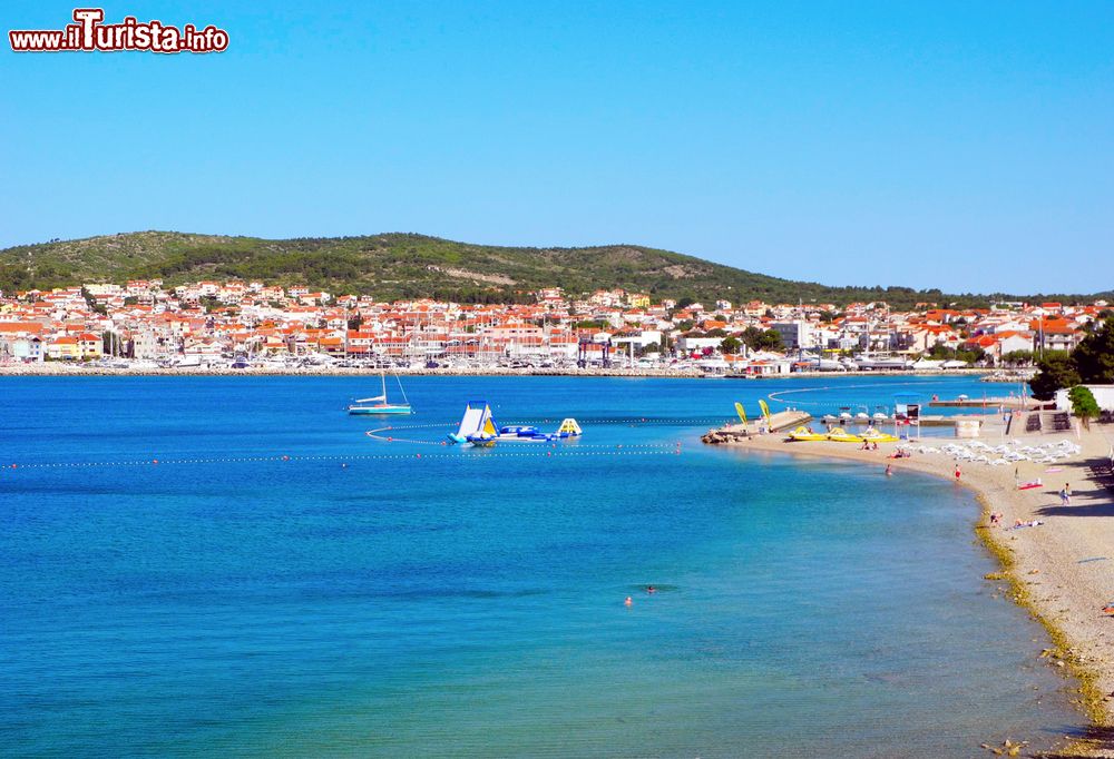 Immagine Una bella spiaggia della città di Vodice con sullo sfondo il centro abitato, Croazia. Vodizze (nome in italiano) si trova lungo la costa, adagiata in una larga baia, a circa dieci km da Sebenico. Le sue coste sono bordate dalle acque cristalline del Mare Adriatico.