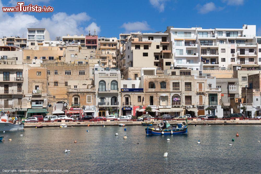 Immagine Uno scorcio della città di Marsascala, isola di Malta, con edifici sullo sfondo. In primo piano, l'acqua del Mediterraneo con barche da pesca - © Yassmin Photo / Shutterstock.com