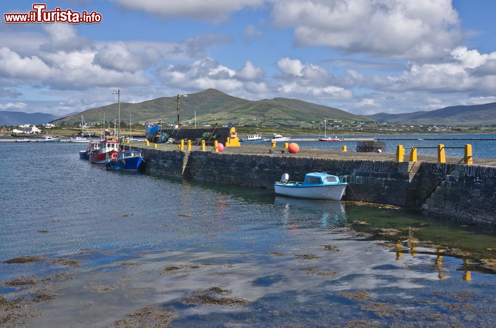 Immagine Il vecchio muro del porto a Knightstown, Valentia Island, Irlanda. Imbarcazioni da diporto e barche da pesca ormeggiate nel porto sono incorniciate da un cielo blu con soffici nuvole che si riflettono nelle acque tranquille del mare.