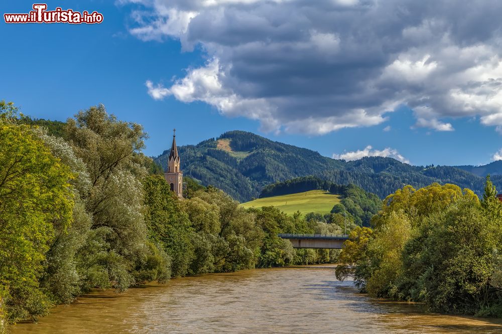 Immagine Veduta del fiume Mura e di una chiesa a Leoben, Austria. A fare da cornice è una natura verde e rigogliosa.