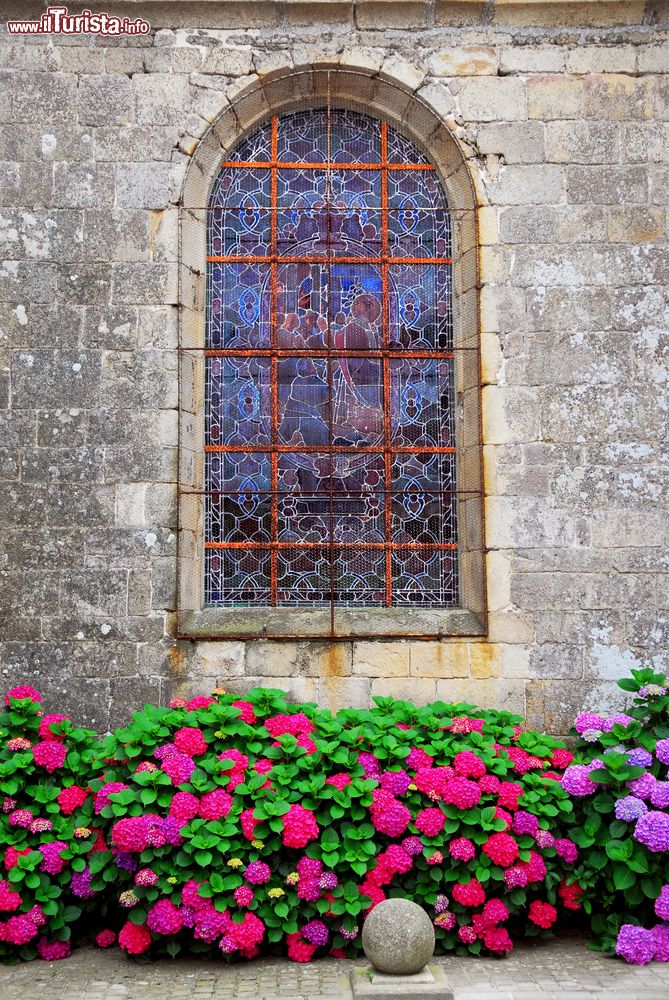 Immagine Vetri colorati e istoriati nella finestra di una chiesa di Carnac, Francia. Ad abbellire l'austero edificio religioso ci sono cespugli di fiori profumati.