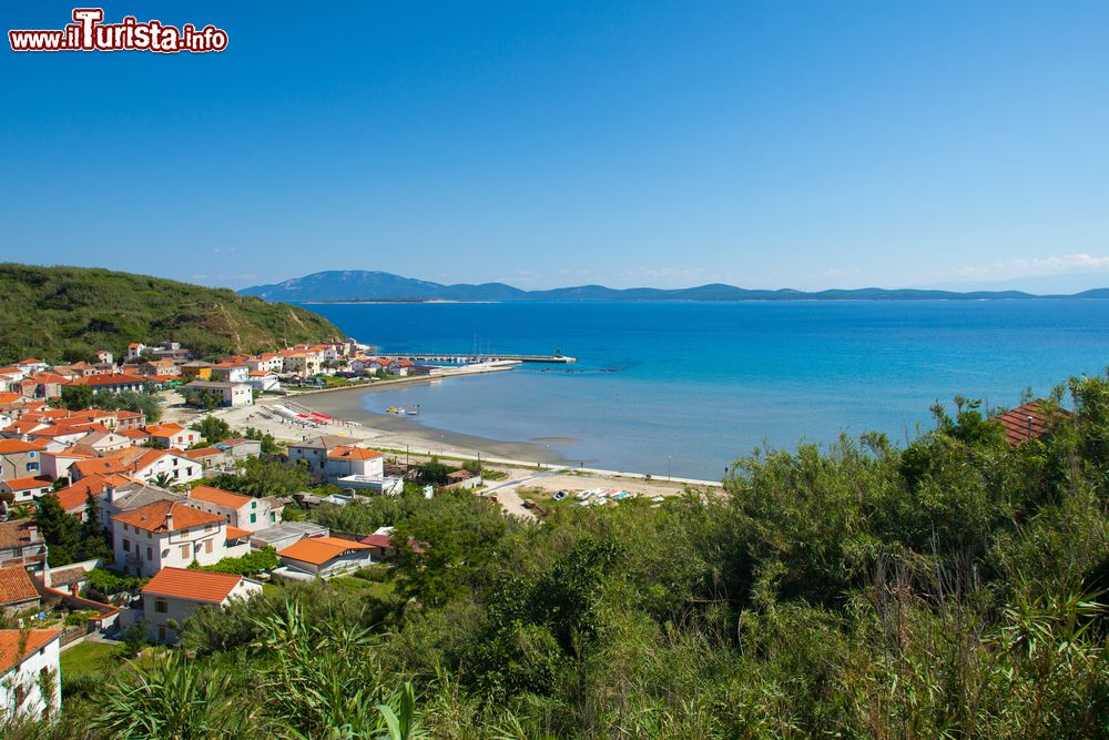 Immagine Vista panoramica della spiaggia principale di Susak (Croazia) e del paese. Sullo sfondo si nota l'isola di Lussino. A Susak non resta che rinfrescarsi nelle fresche acque dell’Adriatico e prendere il sole in una delle tantissime spiagge che la costa propone. Ce n’è per tutti i gusti, la Baia Spiaza ch’è la più estesa dell’isola, attrezzata e ideale per famiglie con bambini, Baia Bok ch’è principalmente una spiaggia nudista, Baia Obis molto frequentata dagli appassionati di pesca subacquea e snorkeling per la sequela di buche, tunnel sottomarini e un canyon a 17 metri di profondità, la rocciosa Baia Nasuzanski, Potarnak, Baia Porat e Punta Valica, quest’ultima spesso in balia di forti venti e onde fragorose.