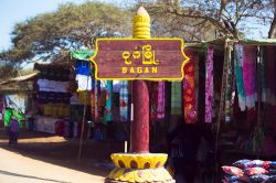 Cartello stradale per Bagan di fronte a un mercato, Myanmar. Stoffe colorate in una bancarella al mercato fanno da sfondo a questo caratteristico cartello di segnalazione stradale che indica ...