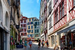 Gente a passeggio in una strada di Bayonne, Francia, in una giornata di sole. Addossate le une alle altre, le case alte di Bayonne con le persiane colorate creano un insieme molto caratteristico ...