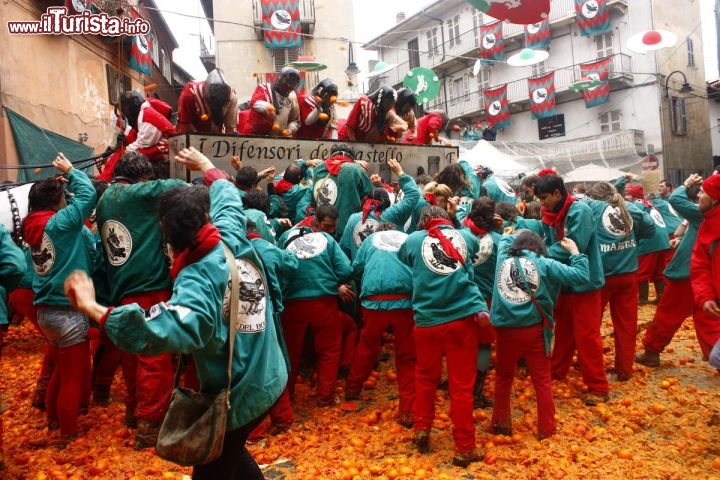 Squadra degli Aranceri Tuchini al Carnevale di Ivrea, durante la scatenata battaglia delle Arance. Questa battaglia sembra abbia avuto inizio verso la metà dell'800, in modo spontaneo con lanci di agrumi a mo' di scherzo gogliardico tra studenti e residenti. Ci sono ben nove squadre a piedi, ciascuna composta da un numero di elementi variabile, comunque qualche centinaio di persone (in totale circa 6.000 persone!), che si scambiano colpi di agrumi con gli aranceri che si trovano sui carri, tirati da coppie di cavalli (pariglie) o da quattro equini (quadriglie). Sono circa 45-50 i carri, ciascuno con una decina/dozzina di aranceri, che indossano protezioni per difendersi dalla furia popolare. Le battaglie si svolgono  nelle varie piazze della centro storico cittadino. Una commisione poi osserva il corso dello svolgimento della battaglia e decreta  le migliori squadre del carnevale - © Archivio della Fondazione dello Storico Carnevale di Ivrea.