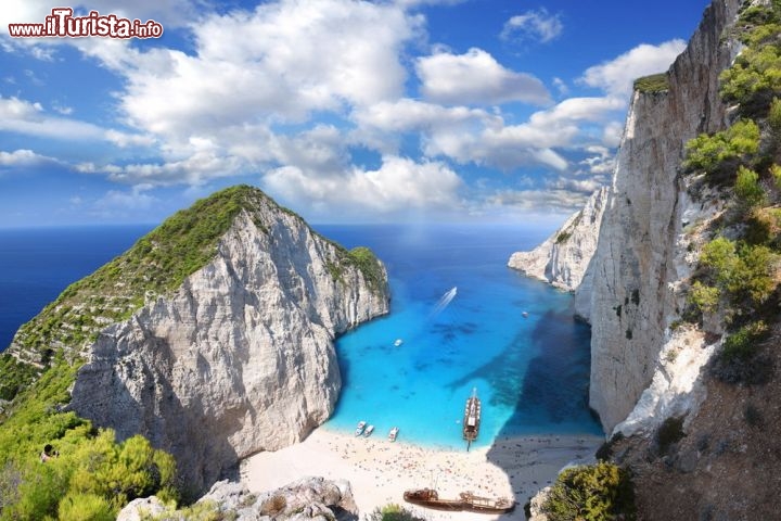 La Spiaggia del Relitto a Zante, Grecia - La spiaggia del Navagio (o del Relitto) a Zante è una delle spiagge più fotografate non solo della Grecia, ma di tutto il mondo. Eppure manca proprio la vostra foto, cosa aspettate? Certamente ne vale la pena. Perché questa selvaggia insenatura greca, raggiungibile unicamente via mare, è uno spettacolo indimenticabile di luci, colori e profumi: la sabbia bianca mista a ciottoli finissimi è abbagliante sotto il sole, e l’acqua, anche grazie alle vicine sorgenti sulfuree, tocca tutte le sfumature immaginabili di azzurri, celesti, turchesi e blu. Abbracciata da alte pareti rocciose, dà il meglio di sé vista dall’alto: sul promontorio in effetti c’è una passerella panoramica sospesa, da cui si possono realizzare scatti da favola. Ovviamente non ci sono servizi né chioschi sulla spiaggia, quindi è bene attrezzarsi con acqua e viveri prima di una gita. Di mattina la Spiaggia del Relitto rimane all’ombra ma ha il vantaggio di essere molto silenziosa, così solitaria da creare un’atmosfera irreale; più tardi arrivano la luce del sole e i turisti, che spesso sbarcano in massa dai battelli delle escursioni. A voi la scelta: meglio la pace o l’abbronzatura?
Il nome della spiaggia è dovuto al relitto dell'MV Panagiotis, arenatosi in questo lido.
Per raggiungerla, se non avete la fortuna di possedere una barca privata, potete noleggiare gommoni o motoscafi o partecipare alle gite di gruppo che partono ogni giorno, a orari diversi, da porto S.Nikolas  - © Samot / Shutterstock.com