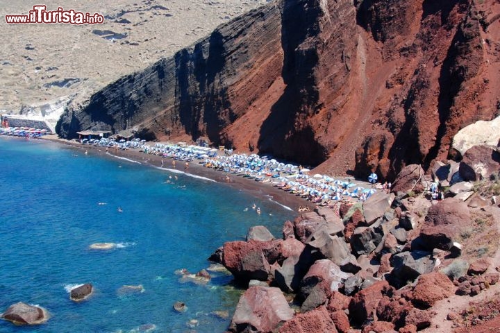 La Spiaggia Rossa di Santorini, Grecia - In una baia a sud-ovest di Santorini, nei pressi di Akrotiri, si apre la Spiaggia Rossa (Kokkini Paralia), sovrastata da un’imponente parete di lava rossastra che contrasta splendidamente con il blu intenso del mare. Incastonata tra gli scogli, è raggiungibile a piedi dal parcheggio in una ventina di minuti, affrontando saliscendi non proprio agevoli, qualche accenno di arrampicata e alcuni punti panoramici - per non dire vertiginosi. Nonostante la posizione inaccessibile, la Spiaggia Rossa è tra le destinazioni più battute di Santorini, e in alta stagione il rischio è che sia troppo affollata per trascorrere una giornata intera di relax. Resta comunque uno splendido scorcio da ammirare e fotografare, con il suo terreno brullo e arso dal sole, i ciuffi di macchia mediterranea, i sassi color mattone e l’acqua limpidissima. Prima di partire, un accorgimento: l’unico chiosco in cui bere e mangiare qualcosa si trova nel parcheggio sovrastante, quindi procuratevi viveri e bevande se intendete restare in spiaggia a lungo. Insomma, se non fosse per la ressa, qualche ombrellone e qualche lettino, sarebbe una vera caletta selvaggia  - © Palis Michalis / Shutterstock.com